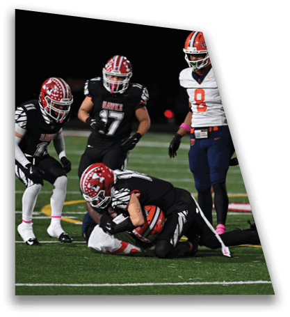Two football players in black uniforms tackle a player in a white and orange uniform on a field while other players watch nearby. Description generated by AI