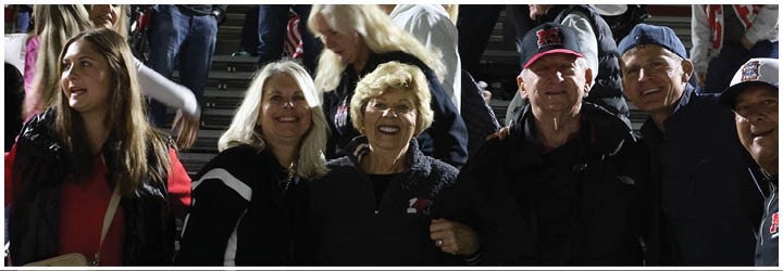 A group of six people standing closely together outdoors at night, smiling and dressed in casual clothing including jackets and hats. Description generated by AI