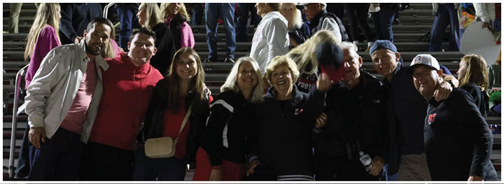 A group of people standing closely together in a stadium setting, smiling and posing for a photo with some wearing casual jackets and hats. Description generated by AI