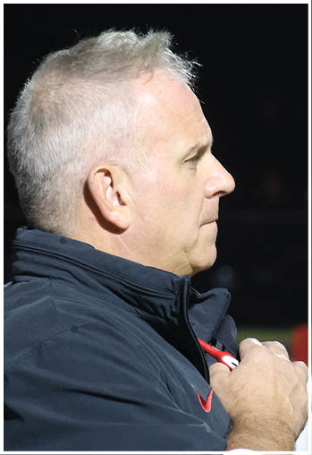 A person with short gray hair wearing a dark jacket is standing outdoors at night, looking to the side with hands clasped near their chest. Description generated by AI