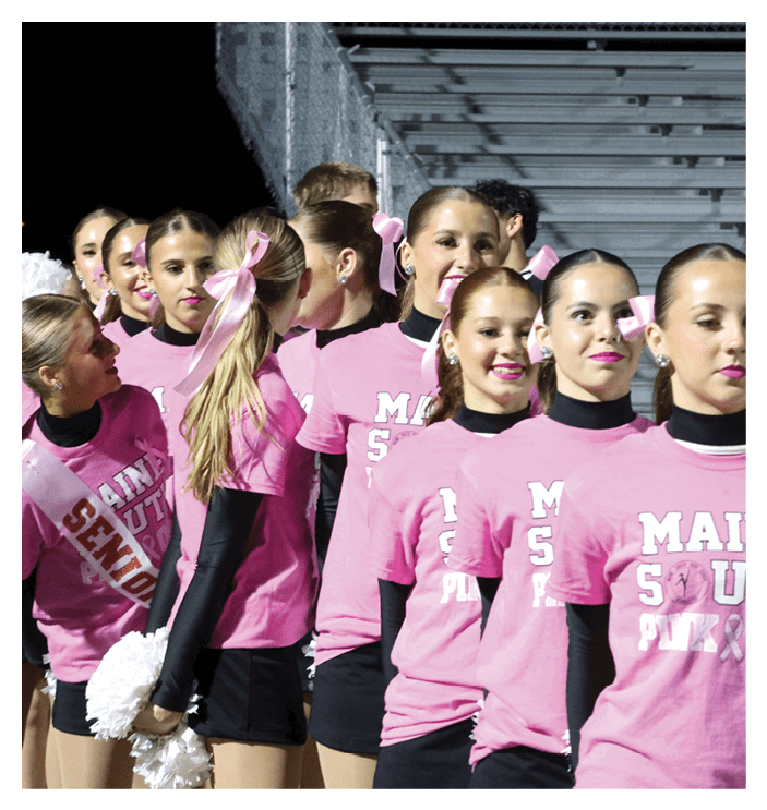 A group of people wearing matching pink shirts and black undershirts stand closely together, some with pink bows in their hair and one holding white pom-poms, appearing to be part of a cheer or spirit team. Description generated by AI