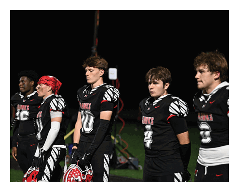 Four people in football uniforms stand on a field at night holding helmets, appearing focused and attentive. Description generated by AI