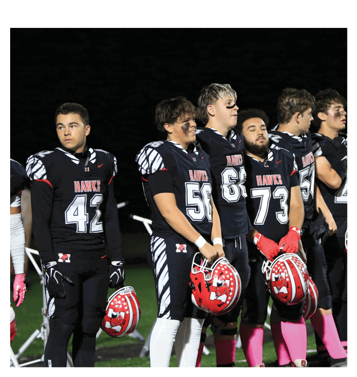 People wearing black football uniforms with pink socks stand in a line on a field at night holding red helmets. Description generated by AI