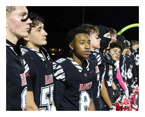 A line of people in black sports uniforms with red and white accents stand side by side on a football field at night, holding red helmets. Description generated by AI