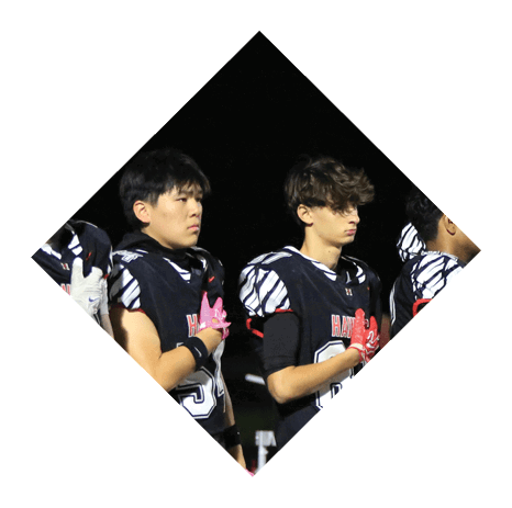 A group of people in black football uniforms with pink gloves and socks stand in a line on a field holding red helmets, appearing to observe a moment of respect. Description generated by AI