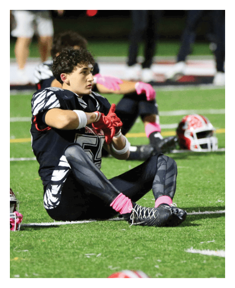 A person in a black and white football uniform with pink gloves and socks is sitting on the field next to a red and white helmet. Description generated by AI