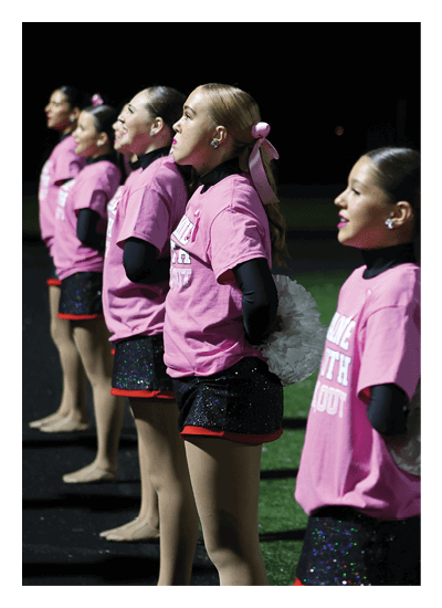 Three people wearing pink shirts and black long sleeves with pink hair ribbons are standing in a row on a field at night, holding white pom-poms. Description generated by AI