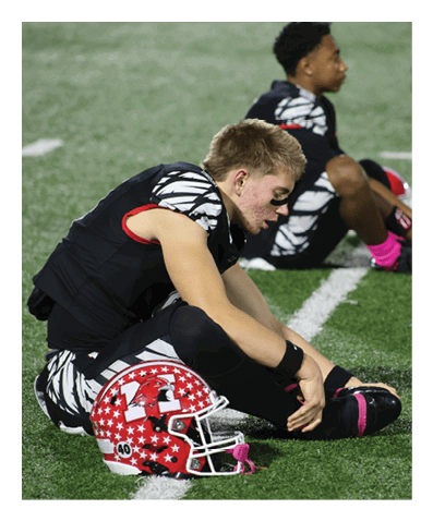 A person in a sports uniform with eye black is sitting on the turf field stretching their leg with a red helmet placed nearby and other players sitting in the background. Description generated by AI