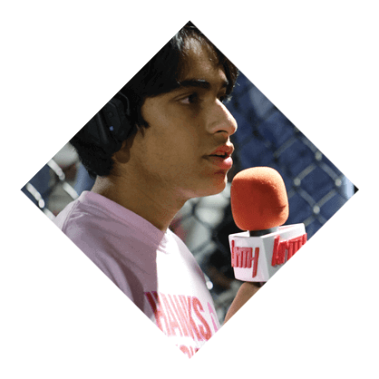 A person wearing a light purple shirt is speaking into a microphone with an orange foam cover and a white label while standing near a chain-link fence. Description generated by AI