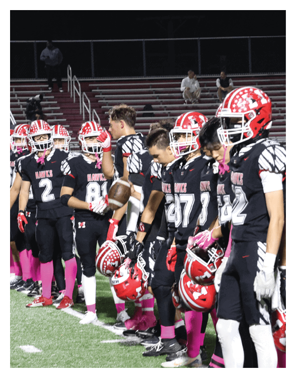 A line of football players in black uniforms with red helmets and pink socks stand on a field at night, some holding helmets and one holding a football. Description generated by AI