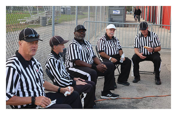 Five adults wearing black and white striped referee shirts and caps are sitting and standing near a chain-link fence and a red building, appearing to take a break. Description generated by AI