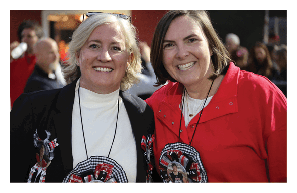 Two adults smiling and standing close together wearing large decorative badges with photos on their clothing in a crowded outdoor setting. Description generated by AI