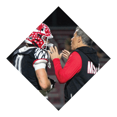 A person wearing a red and white star-patterned football helmet and black and white uniform is standing next to another person in a red long-sleeve shirt who appears to be giving instructions. Description generated by AI