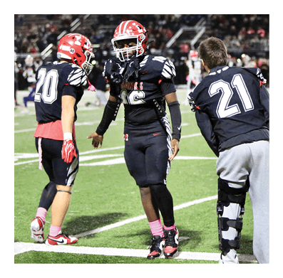 Three people in football uniforms stand on a field, with one person wearing a leg brace and gray sweatpants while the others wear full gear including helmets and pink socks. Description generated by AI