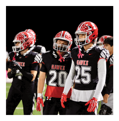 People wearing red and black football uniforms with helmets stand together on a field at night. Description generated by AI