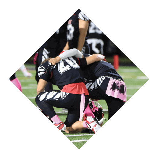 Two people in sports uniforms are on a football field with one person kneeling over the other who is lying on the ground wearing a helmet. Description generated by AI