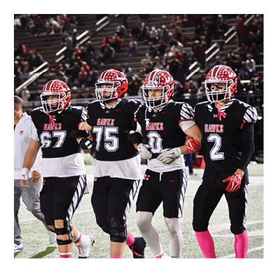 People in football uniforms with pink socks are sitting on a field stretching with their helmets placed on the ground nearby. Description generated by AI
