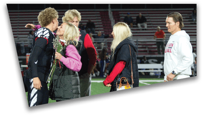 An adult in a sports uniform is being hugged and given flowers by a person in casual clothing on a sports field while two other adults stand nearby watching. Description generated by AI