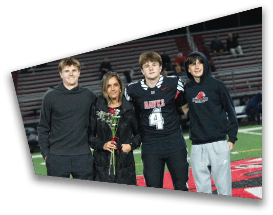 Four people stand together on a football field with one person wearing a football uniform holding flowers. Description generated by AI