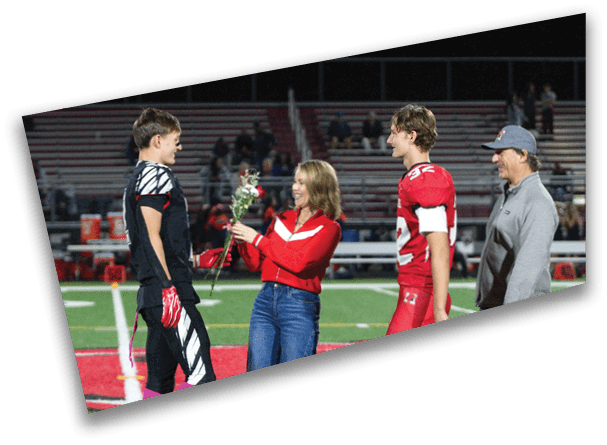 A person in casual clothing is handing flowers to a person in a black sports uniform while another person in a red sports uniform and a person in casual clothing watch on a football field. Description generated by AI