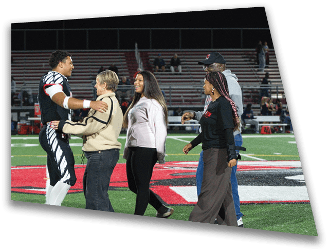 Five people stand on a football field at night with one person in a black and white uniform smiling and interacting with the others who are dressed casually. Description generated by AI