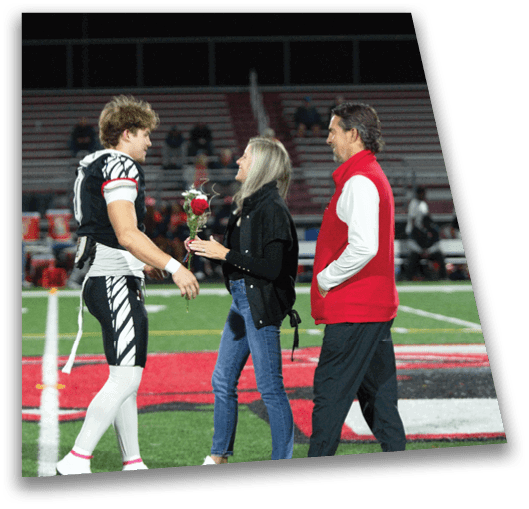 A person in a football uniform is receiving a rose from another person while a third person stands nearby on a football field at night. Description generated by AI