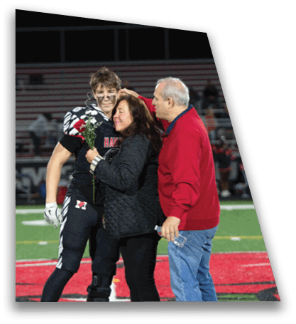 A person in sports gear holding a flower is embraced by another person wearing a black jacket while a third person in a red sweater stands nearby on a sports field. Description generated by AI