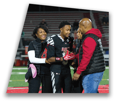 A person wearing a football uniform is smiling and shaking hands with another person dressed in casual clothes on a sports field. Description generated by AI