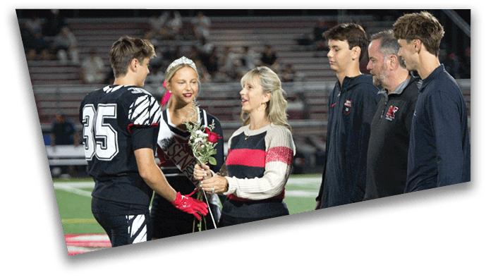A person in a sports uniform is receiving a flower from a person in a striped sweater on a sports field while others stand nearby watching. Description generated by AI