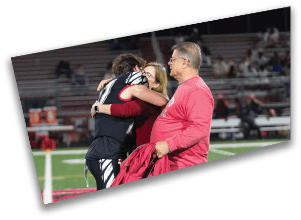 A person in a sports uniform hugs another person wearing glasses and a red sweater on a football field while a third person in a red long-sleeve shirt holds a jacket nearby. Description generated by AI