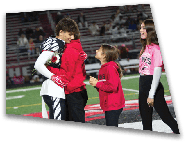 A person in a sports uniform is hugging another person wearing a red hoodie on a football field while a child in a red hoodie looks up at them smiling. Description generated by AI