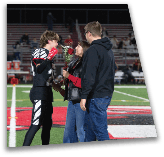 A person in a football uniform is holding a rose and interacting with two people standing on a football field near the end zone. Description generated by AI
