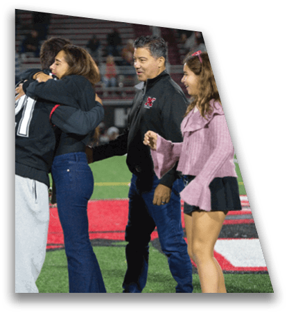 Two people embrace on a sports field while another person stands nearby, with one person wearing a leg brace. Description generated by AI