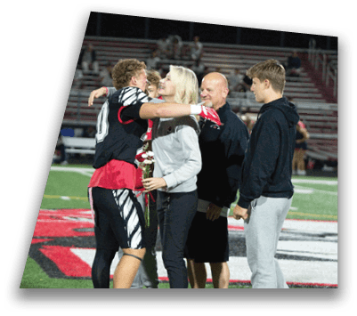 A person in a sports uniform hugs another person holding flowers on a sports field while two other people stand nearby watching. Description generated by AI