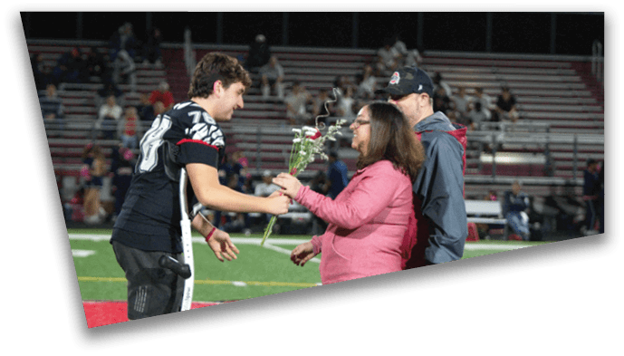 A person with crutches wearing a sports uniform is receiving a flower from another person in casual clothing on a sports field with bleachers in the background. Description generated by AI