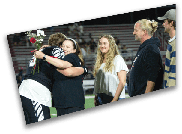 A person in sports attire holding flowers hugs another person while two others stand nearby on a sports field with bleachers in the background. Description generated by AI