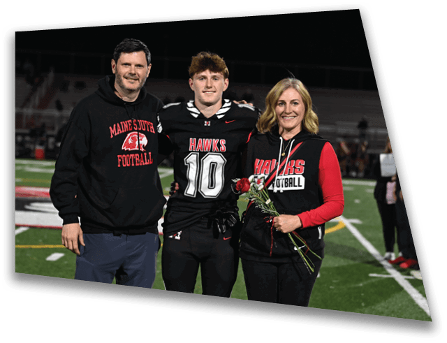 Three people stand on a football field at night with one person in a football uniform holding flowers and the others wearing sports-themed casual clothing. Description generated by AI