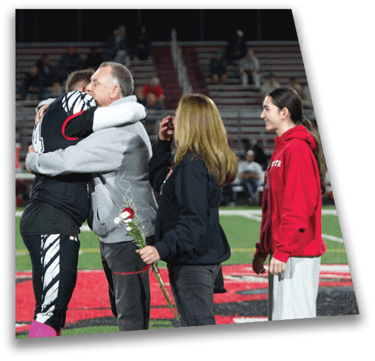 An adult in a sports uniform hugs another adult holding a flower while a third adult stands nearby on a sports field with bleachers in the background. Description generated by AI
