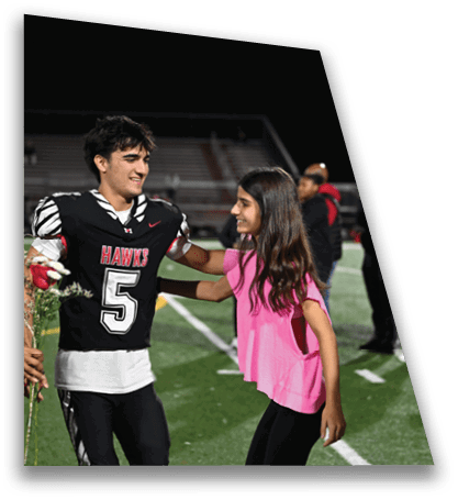 A person in a black football uniform with the number five smiles and embraces another person wearing a pink top on a football field at night. Description generated by AI