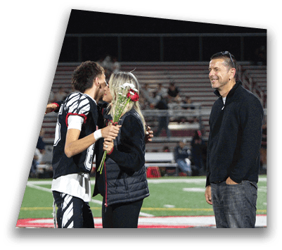 An adult in a sports uniform is holding flowers and embracing another adult on a sports field while a third adult stands nearby smiling. Description generated by AI