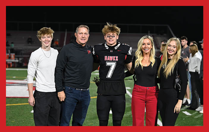 Five people stand together smiling on a football field at night, with one person wearing a black football uniform and the others dressed in casual and semi-formal clothing. Description generated by AI