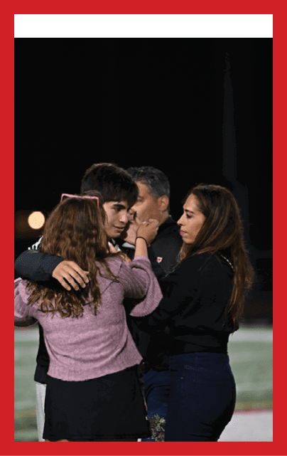 A person in a football uniform with the number 52 stands on a field at night smiling with two adults on either side, one holding flowers. Description generated by AI