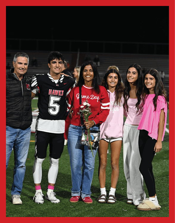 A person in a black and white sports uniform with the number 74 is smiling and talking to two other people, one wearing a dark jacket and the other wearing a red top, on a sports field at night. Description generated by AI