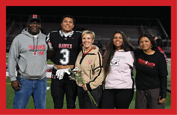 Five people stand together on a football field at night with one person in a football uniform holding flowers and the others wearing casual clothing. Description generated by AI