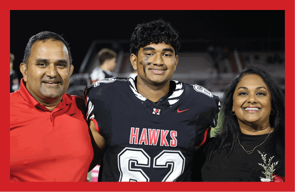 A person wearing a black football uniform with the number 62 and the word “HAWKS" stands between two smiling adults, all posing together outdoors at night. Description generated by AI