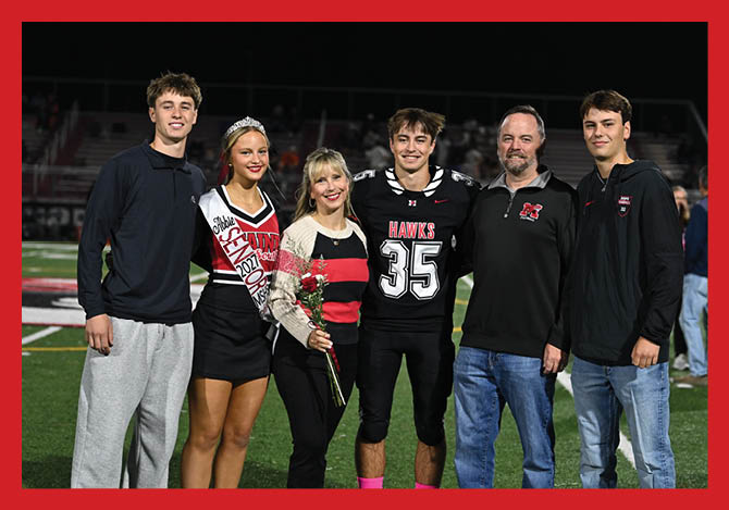 Six people stand together on a sports field at night with one person wearing a football uniform, another wearing a sash and holding flowers, and the others dressed casually. Description generated by AI