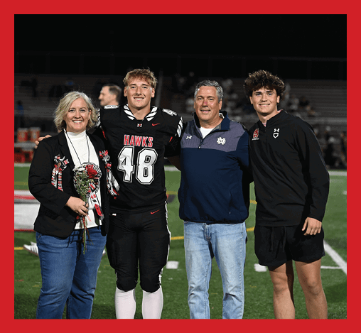 Four people stand together on a sports field at night with one person wearing a football uniform holding flowers. Description generated by AI