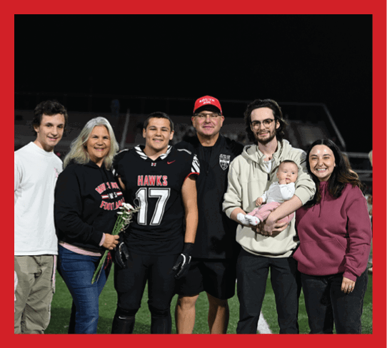 A group of six adults and one newborn stand together on a sports field at night, with one adult wearing a football uniform and others dressed casually, smiling and posing for the photo. Description generated by AI
