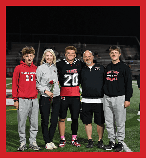 Five people stand together on a football field at night with one person wearing a football uniform and others dressed casually, while some hold flowers. Description generated by AI
