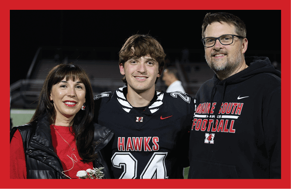 Three people smiling on a sports field at night with one person wearing a football uniform and the others wearing casual clothing with team logos. Description generated by AI